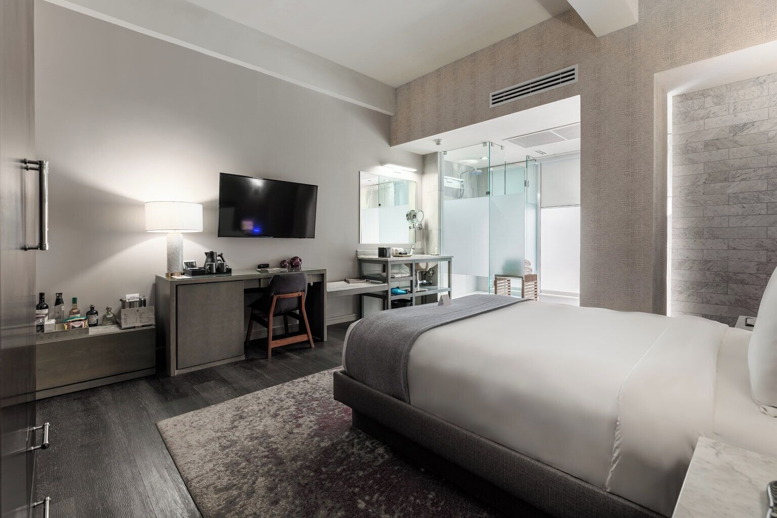 A modern hotel room with a bed, desk, TV, and a glass shower area.
