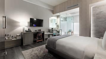 A modern hotel room with a bed, desk, TV, and a glass shower area.