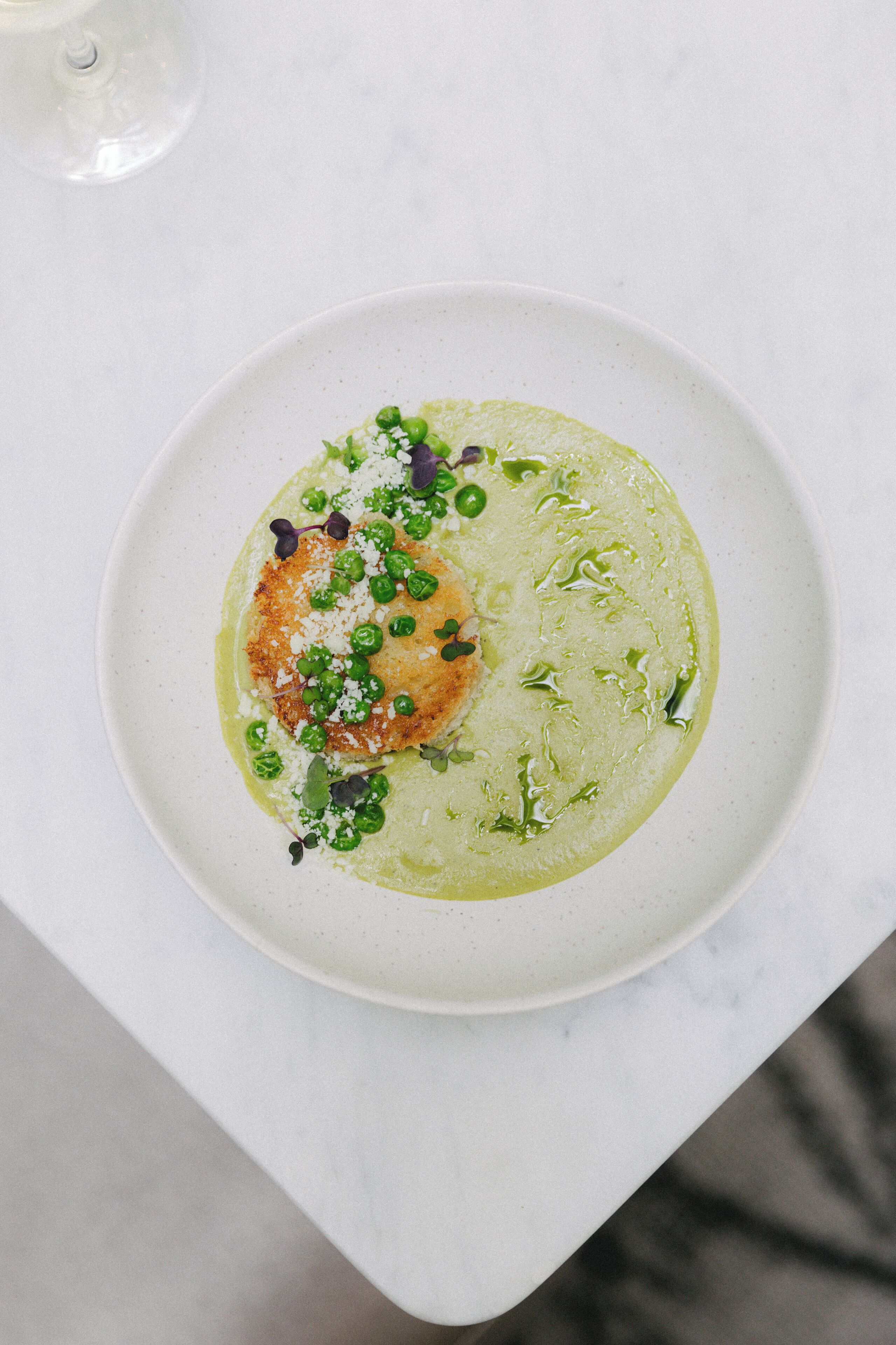A white plate with a green sauce, green peas, and a piece of food on a white table.