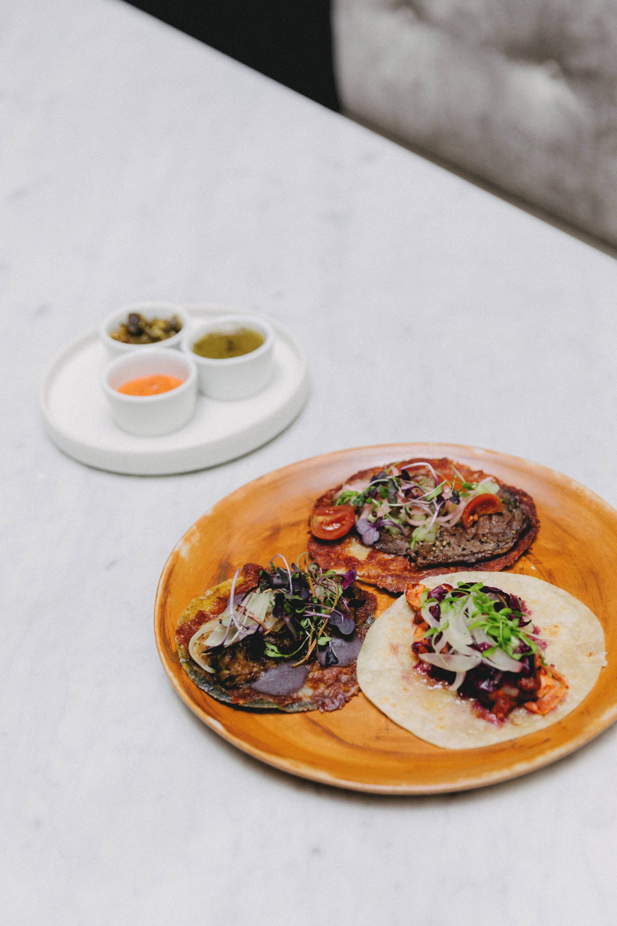 Un plato con tres tacos y dos recipientes pequeños de salsas sobre una mesa blanca.