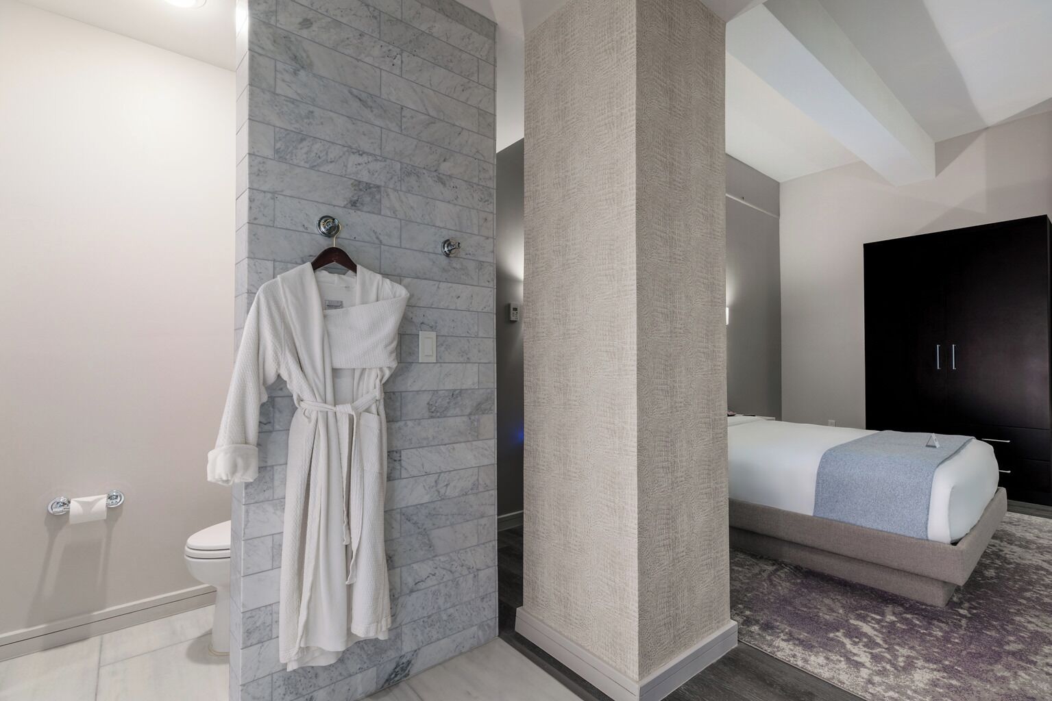 A modern bathroom with a marble wall, a white bathrobe hanging, a toilet, and a bed with a blue blanket.