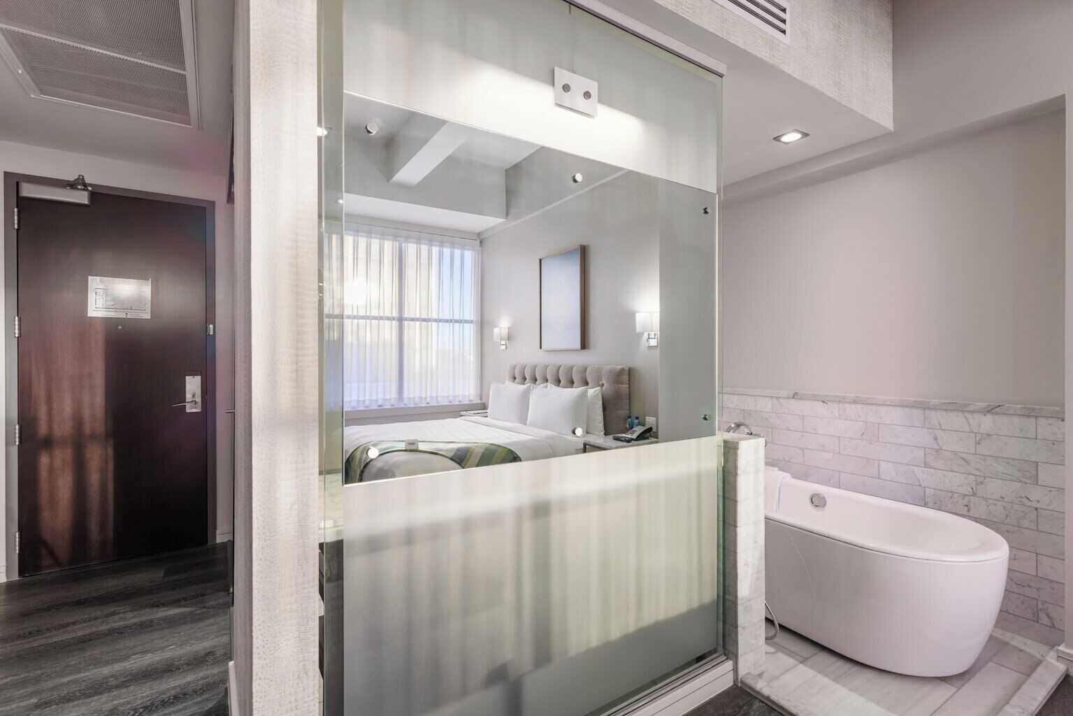 A modern hotel room with a glass partition, a bed, a nightstand, a bathroom with a bathtub, and a window with curtains.