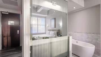 A modern hotel room with a glass partition, a bed, a nightstand, a bathroom with a bathtub, and a window with curtains.