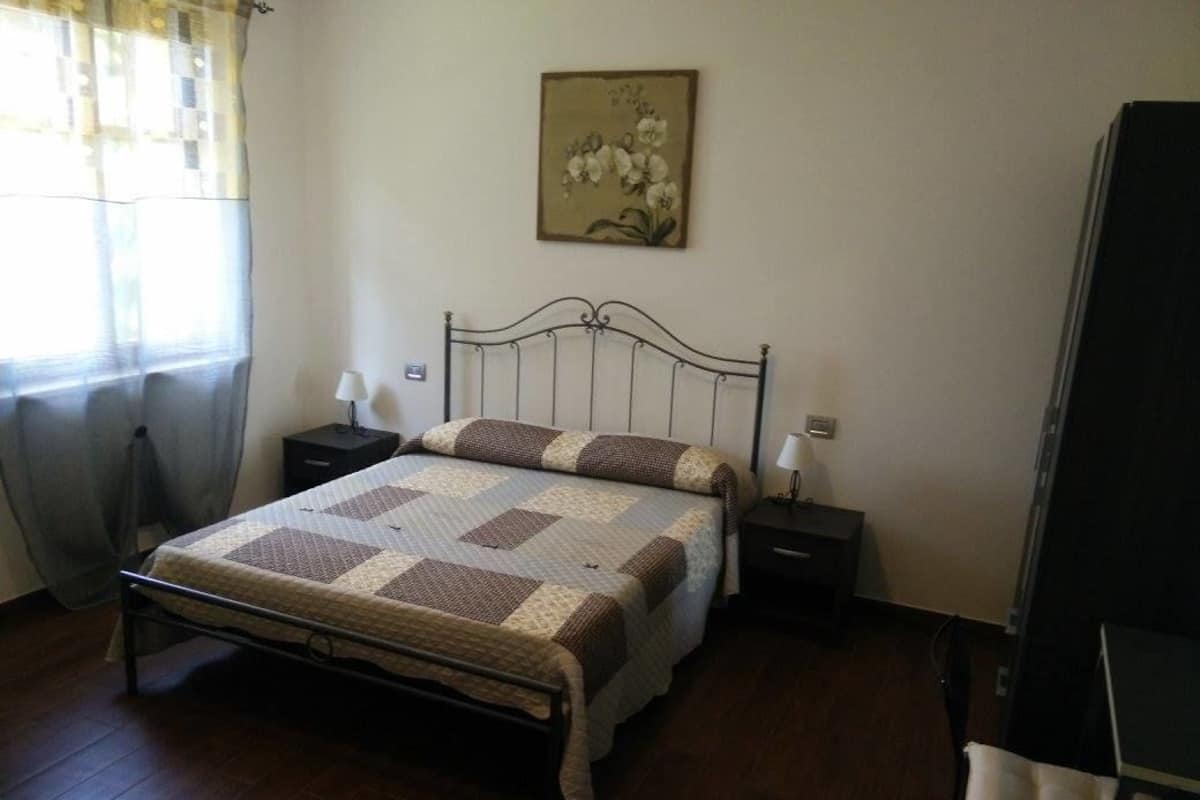 A bedroom with a bed, two bedside tables, a nightstand, a chair, a window with curtains, and a wall-mounted picture.