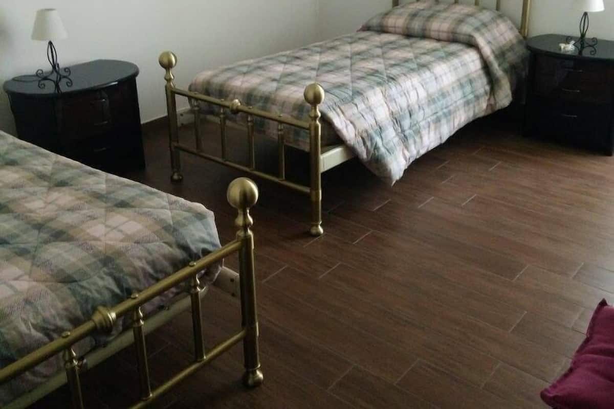 A room with two beds, a wooden floor, a painting on the wall, and a nightstand with a lamp.