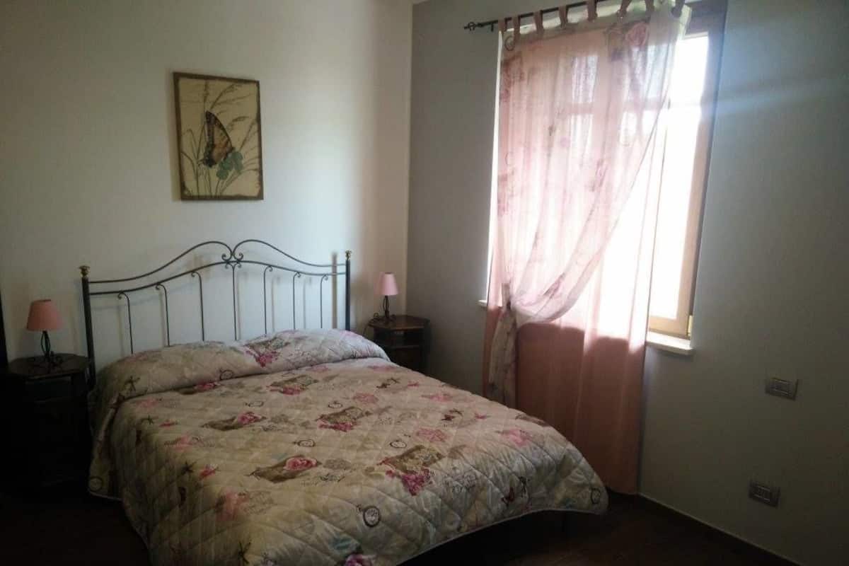 A bedroom with a bed, a nightstand, a lamp, a window with curtains, and a wall-mounted picture.