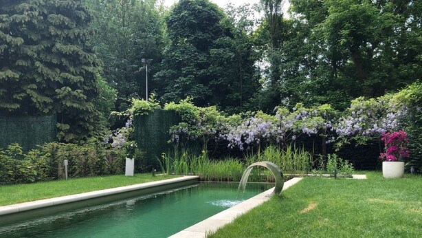 A rectangular pool with a curved water feature, surrounded by lush greenery and flowering plants. - Exclusive Flats Studio Swimming-pool (Dilbeek)
