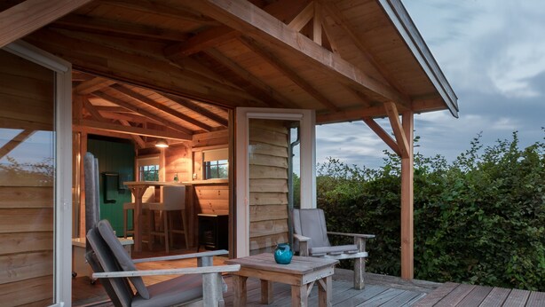 A wooden deck with a covered seating area, a table, and a chair. - Aan de Waterspiegel (Aagtekerke)