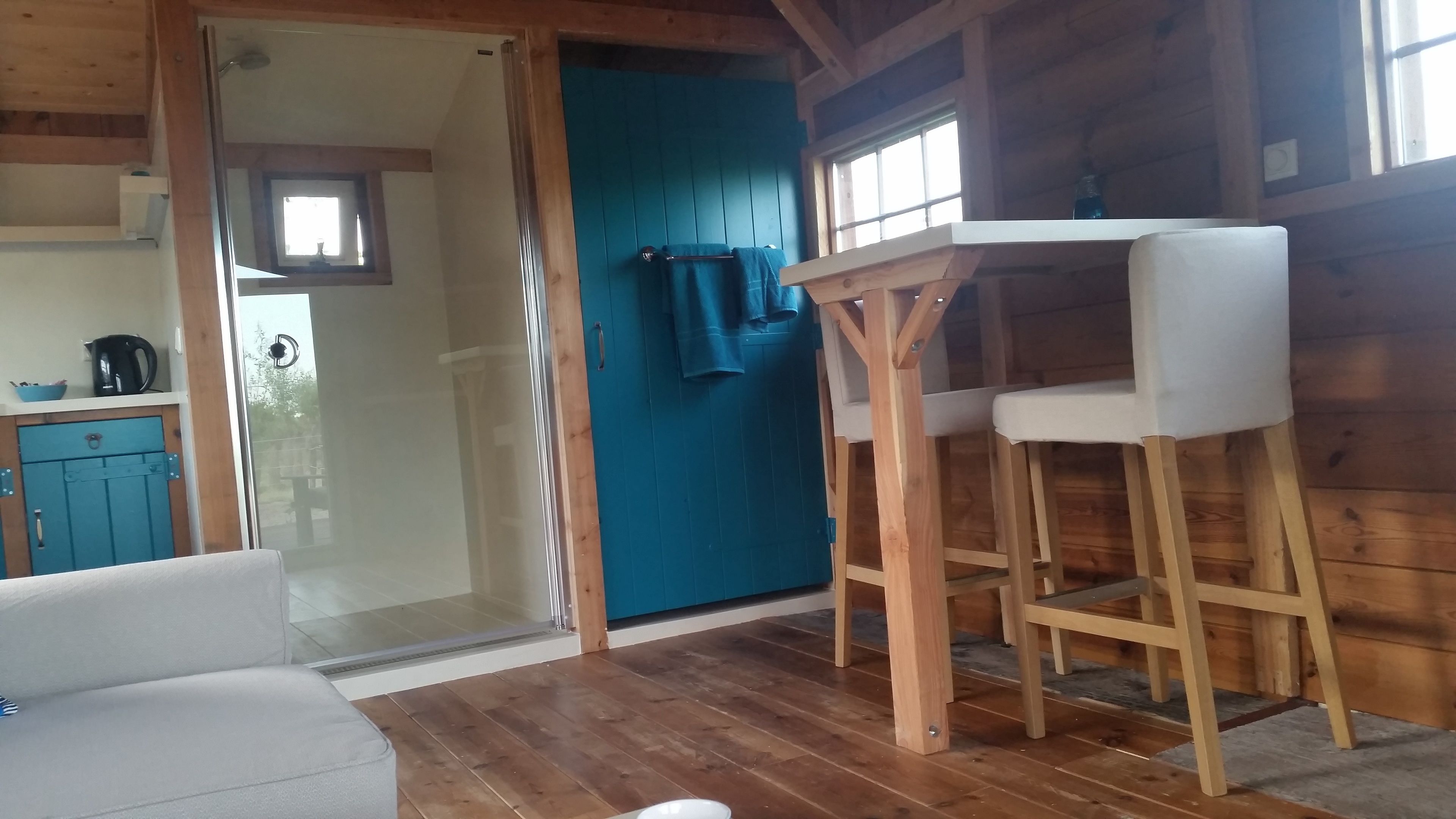 Een gezellig houten interieur met een houten vloer, een witte bank, een houten eettafel met twee stoelen, een blauwe deur en een zichtbare keuken door een glazen deur.