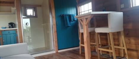 Een gezellig houten interieur met een houten vloer, een witte bank, een houten eettafel met twee stoelen, een blauwe deur en een zichtbare keuken door een glazen deur.