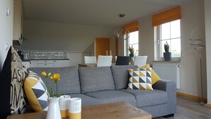 A modern living room with a grey sofa, wooden coffee table, and decorative vases. A kitchen is visible in the background with white cabinets and a window with orange blinds. - Aan de Waterspiegel (Aagtekerke)