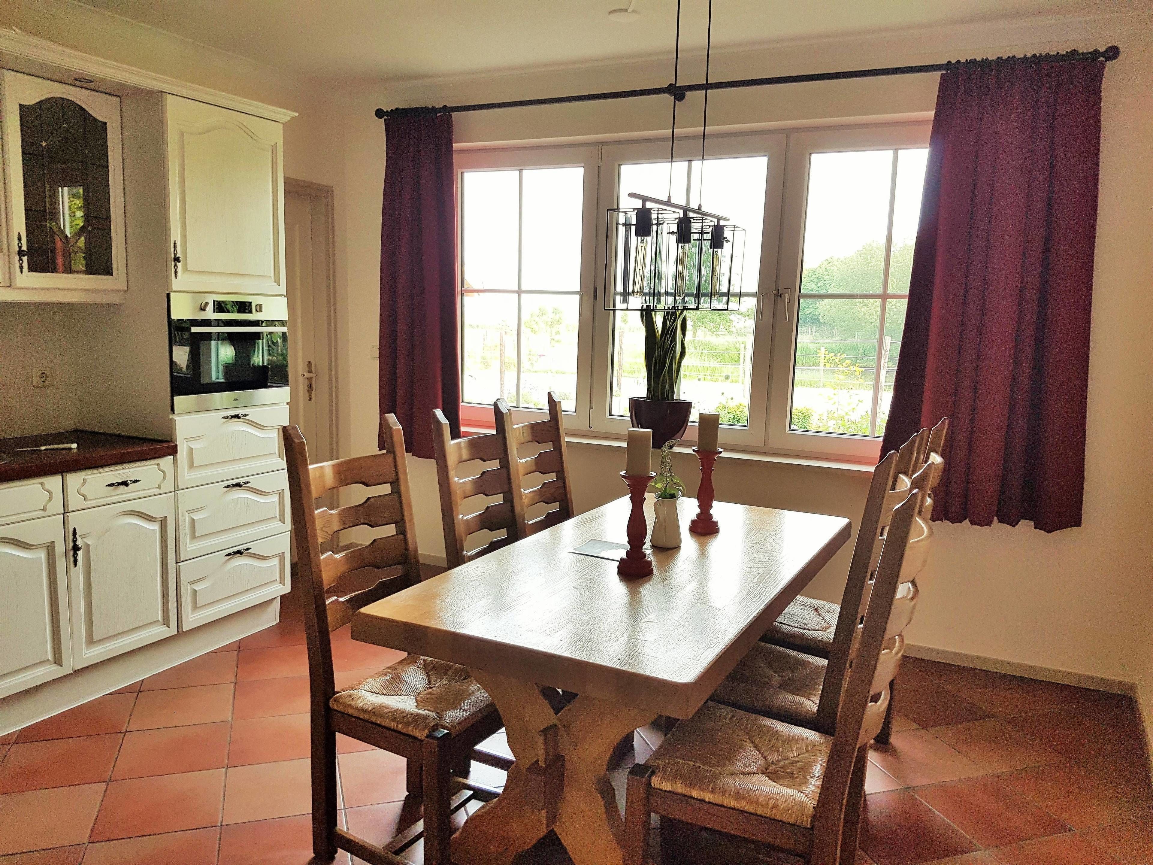 A dining area with a wooden table and chairs, a kitchen with white cabinets, and large windows with curtains.