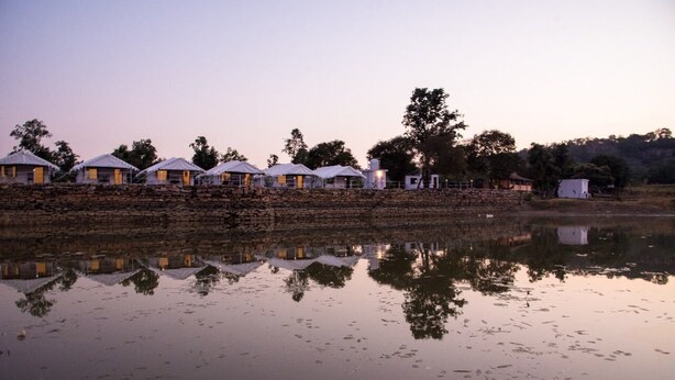 A row of white-roofed cottages along a stone wall by a calm body of water, reflecting the structures and trees.