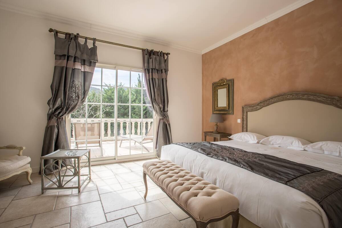 A bedroom with a large bed, a bench, a small table, and a balcony with a chair.