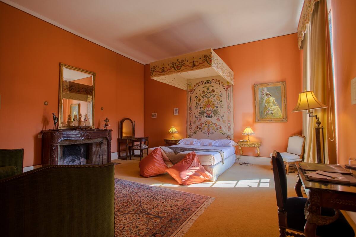 A bedroom with a large bed, a fireplace, a mirror, and a painting on the wall.