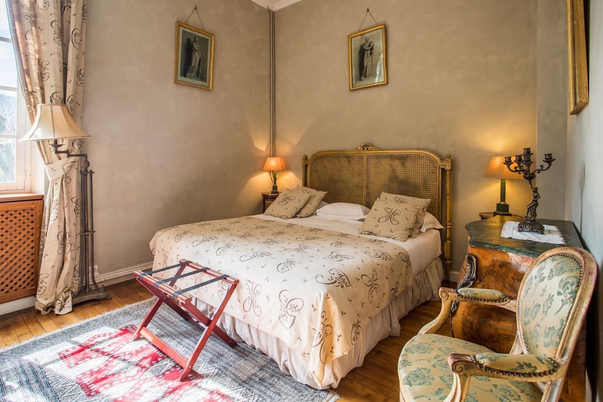 A bedroom with a bed, two bedside lamps, a chair, a small table, and a window with curtains.