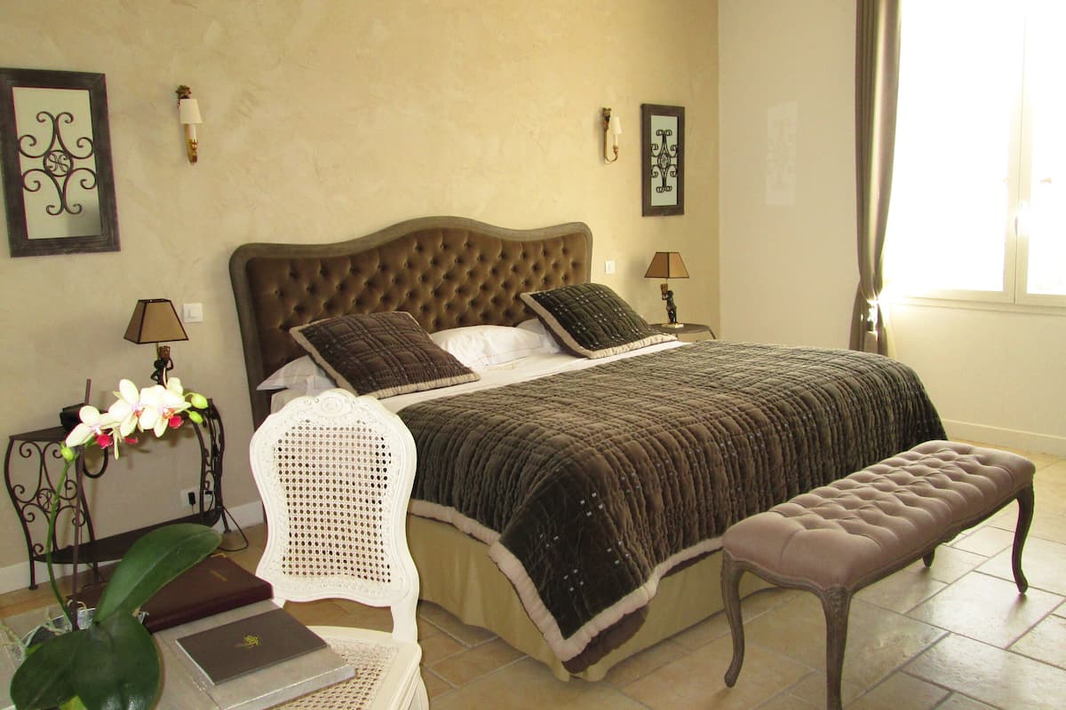 A bedroom with a tufted headboard, a large bed with a dark quilt, a bedside table with a lamp, a chair, and a bench.