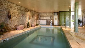 An indoor swimming pool with stone walls, a wooden bench, and a statue. - Hôtellerie du Château de Floure & Spa (Floure)