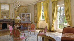 A room with a fireplace, a chandelier, a bookshelf, and a large window with curtains. - Hôtellerie du Château de Floure & Spa (Floure)