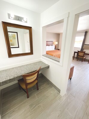 Quarto de hotel com bancada de granito, cadeira de madeira, cama com colcha laranja e um espelho grande.