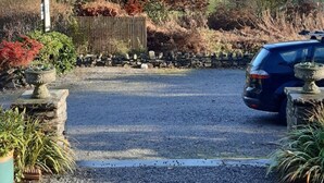 Une allée de gravier mène à une voiture garée, le tout étant entouré d’un jardin où poussent différentes plantes et d’une clôture en bois.