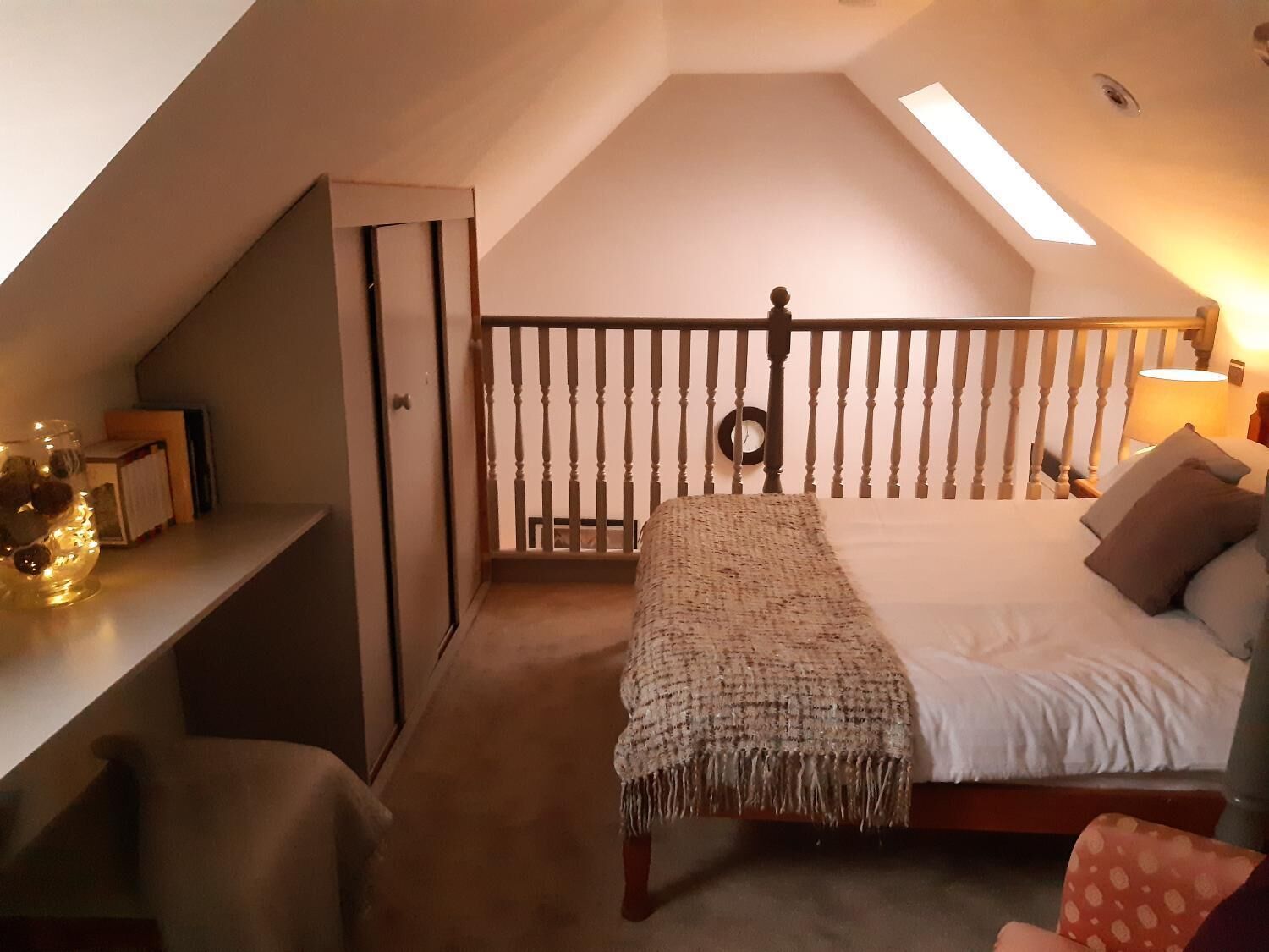 Un dormitorio acogedor en el ático, con techo inclinado, cama de madera con manta tejida, un escritorio con lámpara y una ventana pequeña.