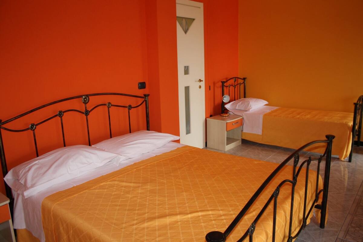 Een kamer met twee bedden, elk met een metalen frame en oranje beddengoed. Er is een witte deur en een klein nachtkastje met een lamp.