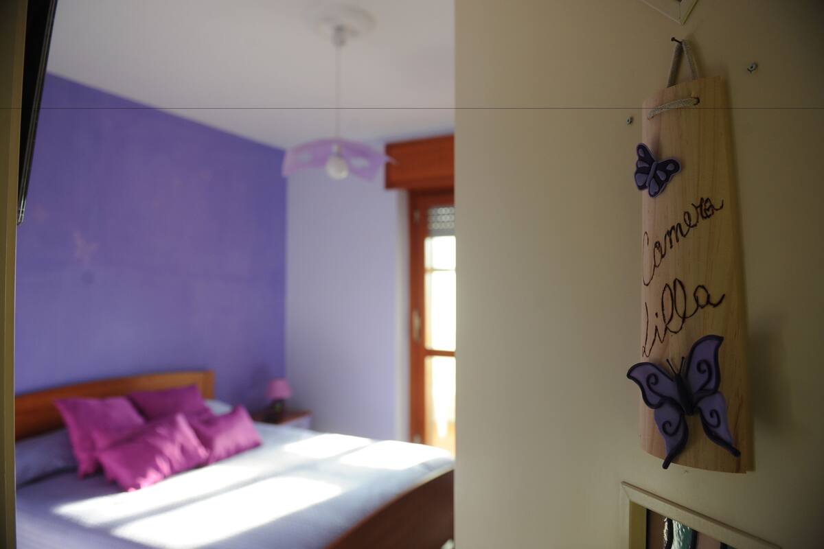 Een slaapkamer met een bed, paarse wanden, een houten hoofdeinde en een bordje aan de muur met een vlinder en tekst.