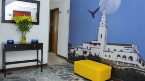A lobby with a stone floor, a yellow bench, a vase with yellow flowers, a blue wall with a mural of a building and moon, and a mirror on the wall. - Hotel Normandia Gold (Bogotá)