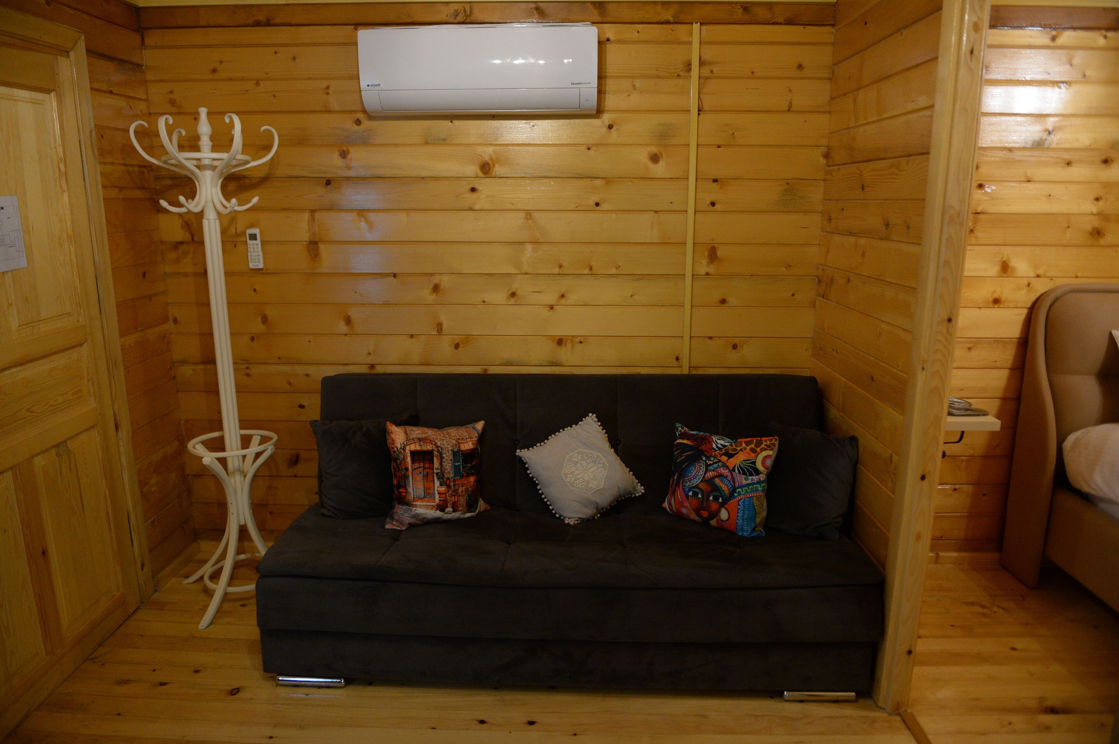 Interior de una cabaña de madera con un sofá negro, tres almohadas, un perchero blanco y un aire acondicionado montado en la pared.