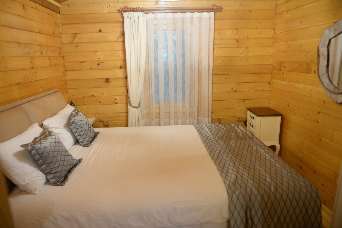 Un dormitorio de cabaña de madera con cama, almohadas, mesita de noche y ventana con cortinas.