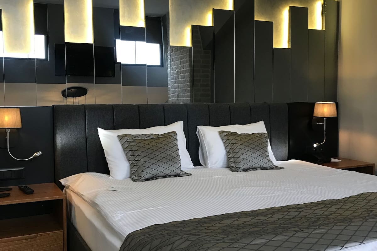 Una habitación de hotel moderna con una cama grande, dos mesitas de noche, un televisor montado en la pared y una lámpara con un diseño geométrico original.