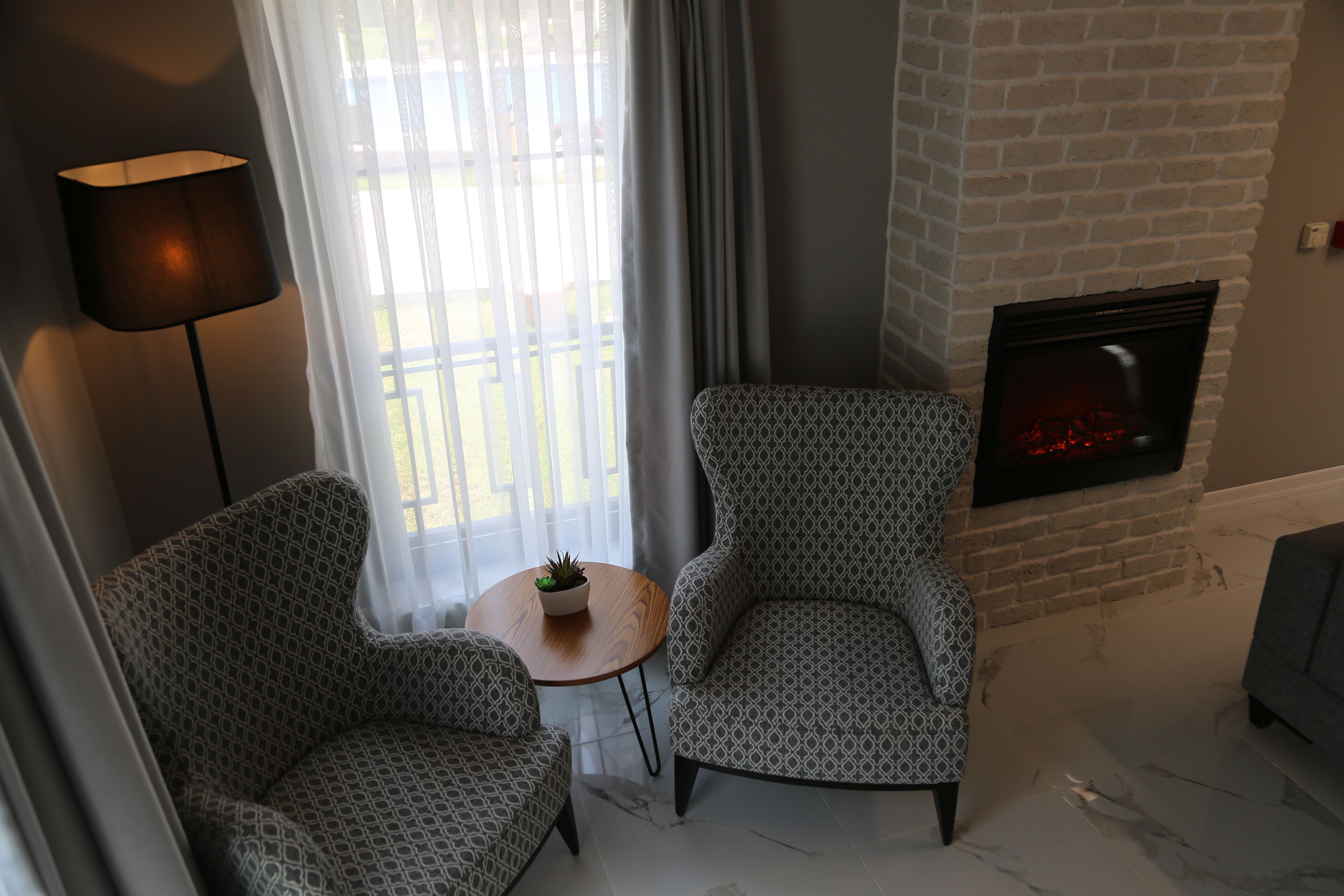 Een moderne woonkamer met twee gestreept fauteuils, een kleine ronde tafel met een plant, een open haard en uitzicht door een raam met gordijnen.