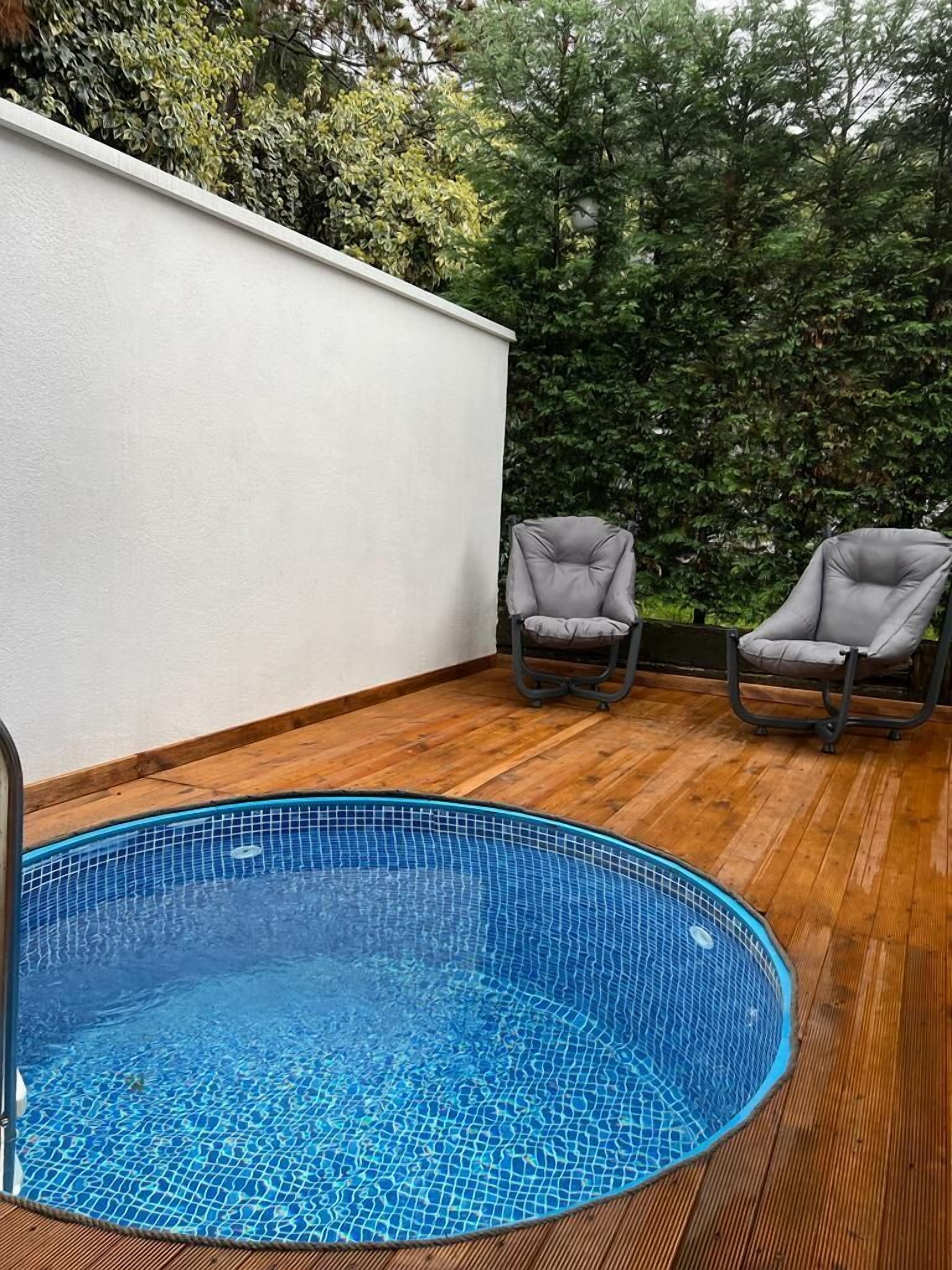A small, round, blue-tiled pool with a wooden deck and two grey cushioned chairs.