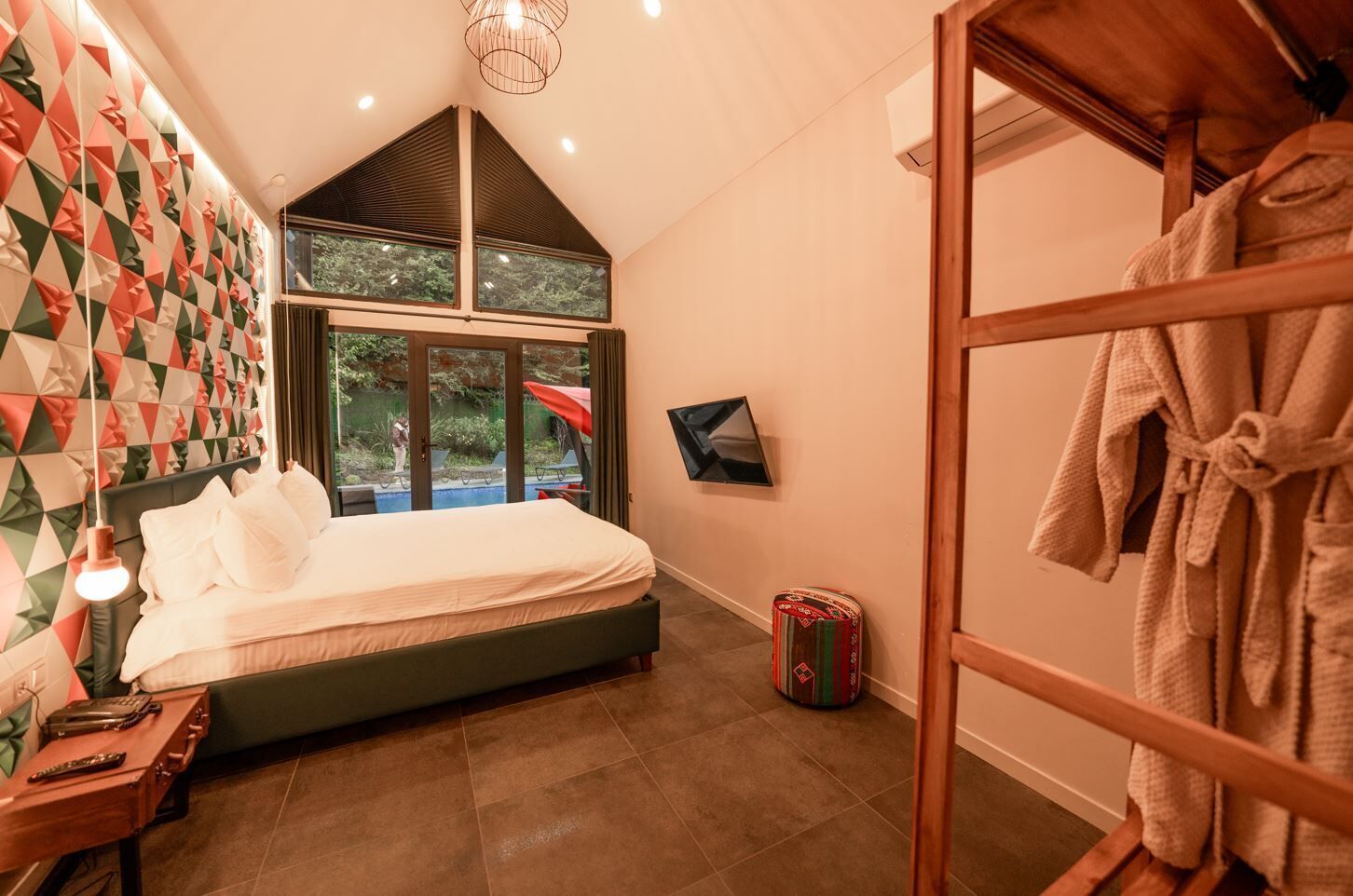 Un dormitorio con cama, mesita de noche, lámpara, ventana grande y vista a una piscina.