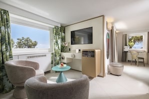 A modern hotel room with a large window, a flat-screen TV, a small kitchenette, and two comfortable armchairs.