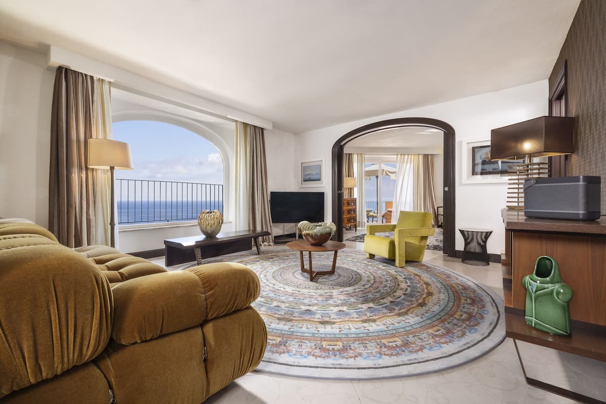 A spacious living room with a large window, a round rug, a sofa, a television, and a green handbag on a wooden table.