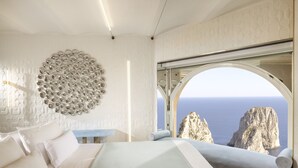 A bedroom with a bed, a wall art installation, and a large window offering a view of the sea and cliffs. - Hotel Punta Tragara (Capri)