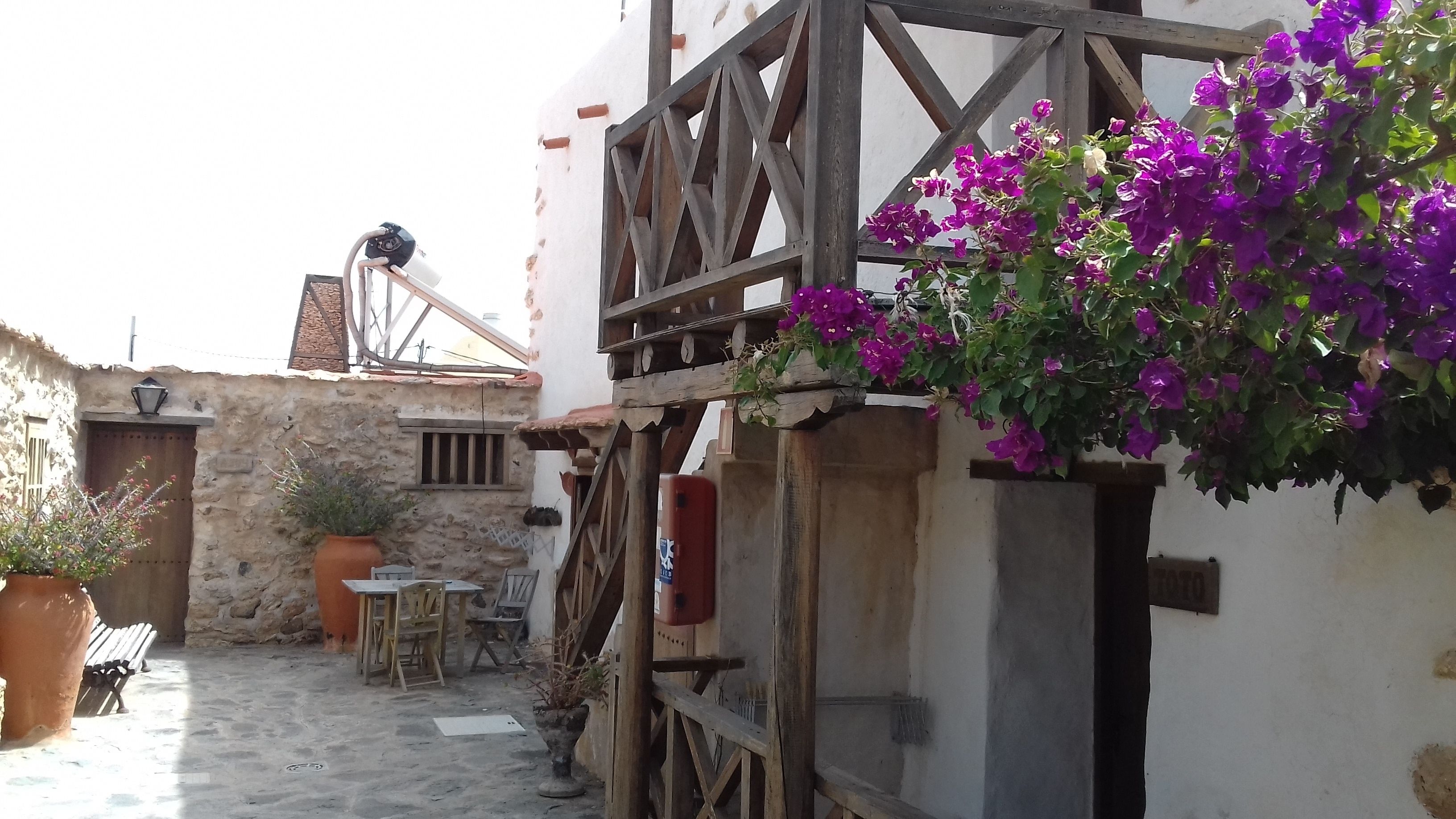 Une cour rustique avec un mur de pierre, des meubles en bois et des fleurs violettes suspendues.