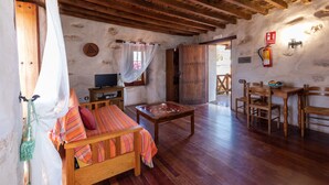 Une pièce chaleureuse avec un canapé en bois, une table en bois et une table à manger avec des chaises. On y trouve un téléviseur posé sur un meuble en bois, un extincteur accroché au mur et une fenêtre avec des rideaux.