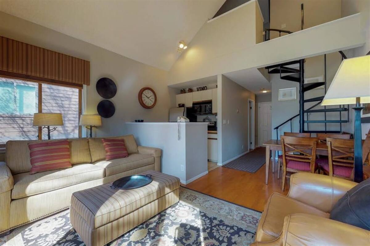 A spacious living area with a sofa, ottoman, and a dining table with chairs. A kitchen is visible in the background, and there is a staircase leading to an upper level.