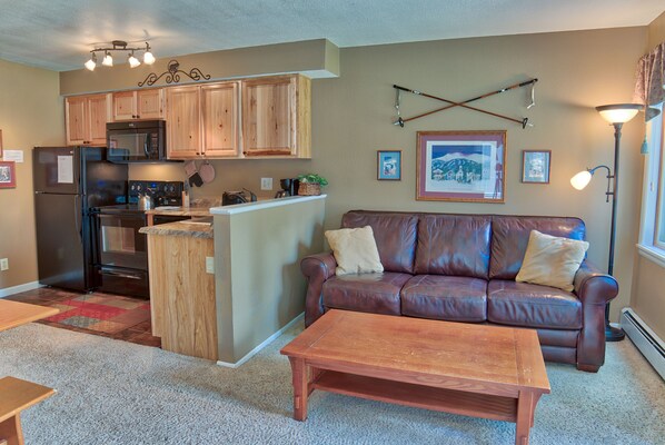 A kitchen with wooden cabinets, a refrigerator, and a microwave. There is a brown leather couch with cushions and a wooden coffee table.