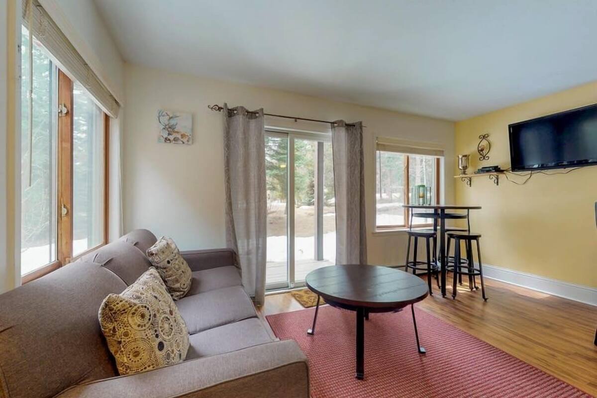 A living room with a sofa, a small round table, a TV mounted on the wall, and a sliding glass door leading to an outdoor area.