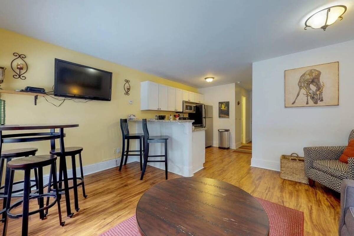 A living room with a round wooden table, a TV mounted on the wall, and a kitchen area with white cabinets and a microwave.