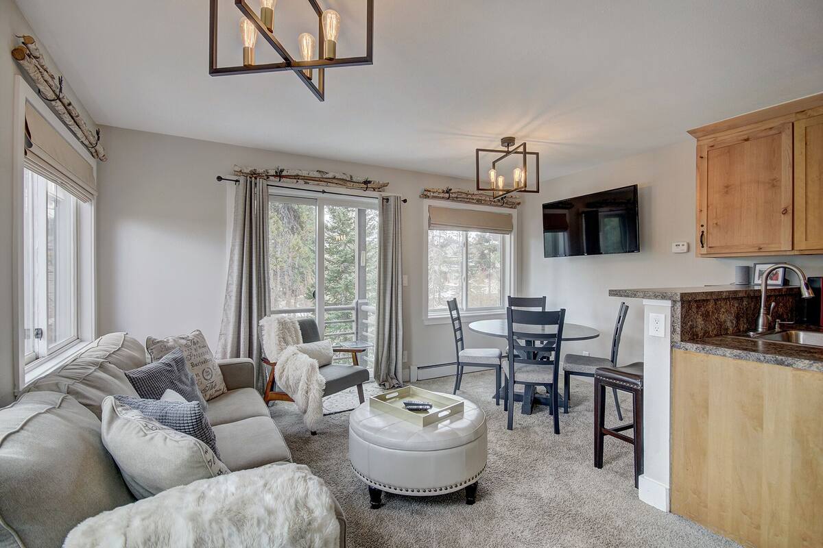 A cozy living room with a sofa, ottoman, and chairs, a dining area with a table and chairs, and a kitchen with wooden cabinets and a TV.
