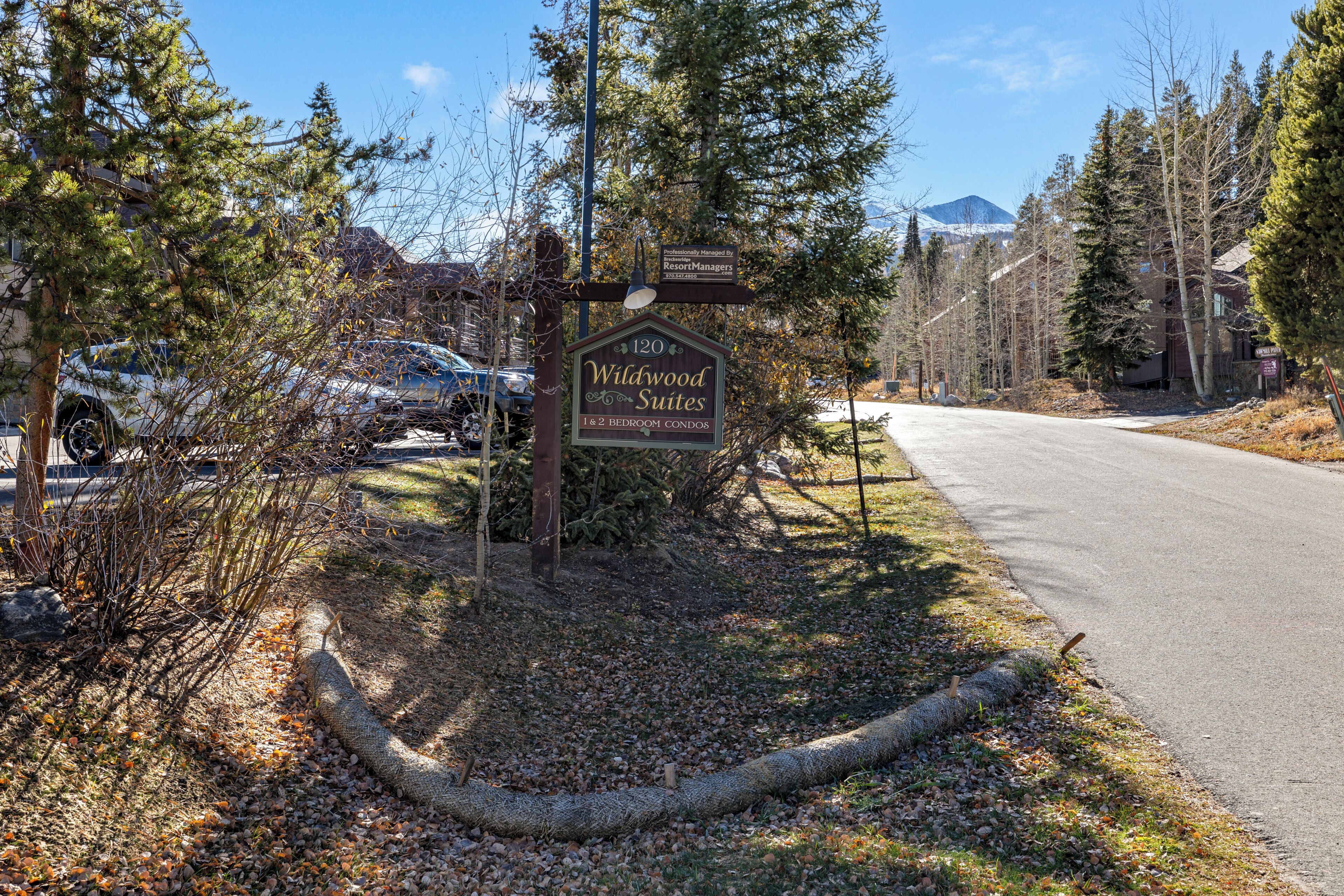 A sign for Wildwood Suites indicating 1 & 2 bedroom condos, a paved road, and surrounding trees.