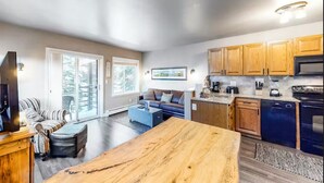 A spacious living area with a large wooden table, a sofa, and a television. There is a kitchen area with wooden cabinets and a dishwasher. - Wildwood Lodge Downtown Condos with Hot Tub (Breckenridge)