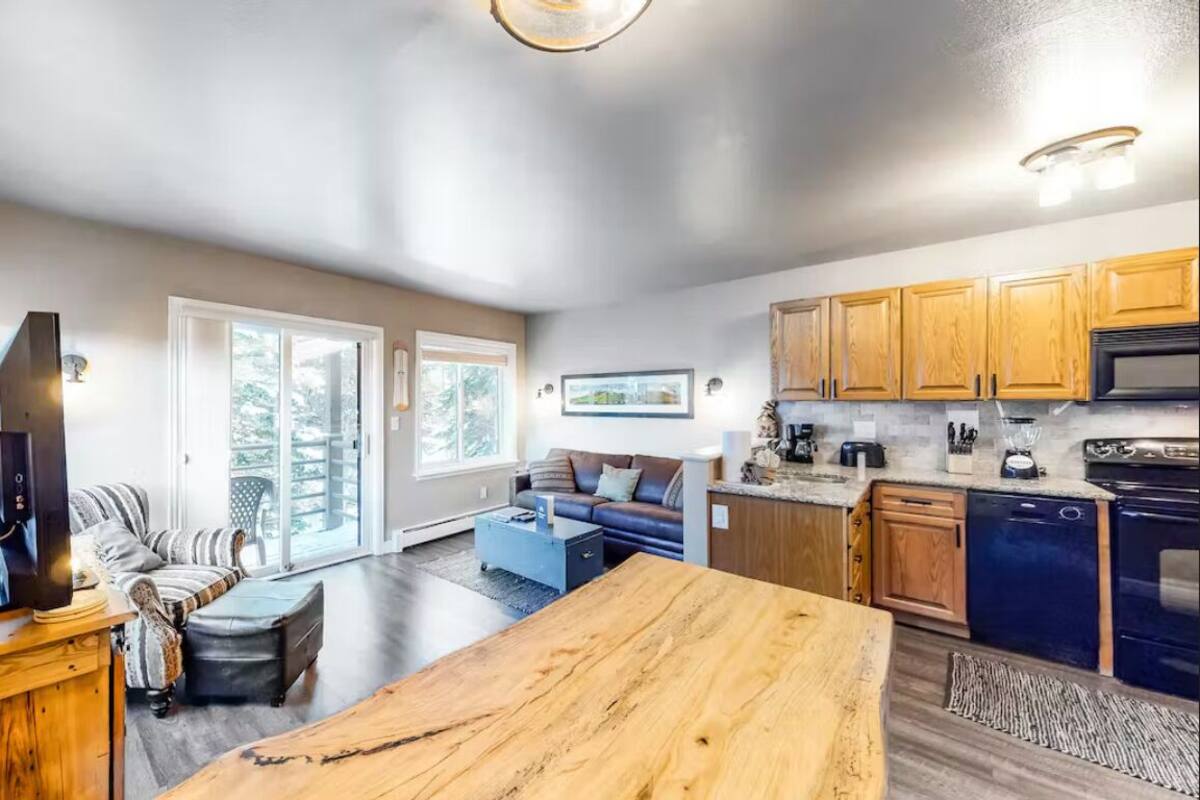 A spacious living area with a large wooden table, a sofa, and a television. There is a kitchen area with wooden cabinets and a dishwasher.