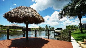 A wooden deck with a thatched umbrella, a water body, and a palm tree. - Bahama by 123cape VR (Cape Coral)