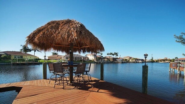 A wooden deck with a thatched umbrella and chairs overlooking a calm body of water, with buildings in the background.